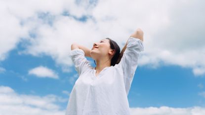 how to work on self love: Low angle portrait of relaxed young woman with her eyes closed, arms outstretched around fresh air and sunlight with head looking up to the blue sky.