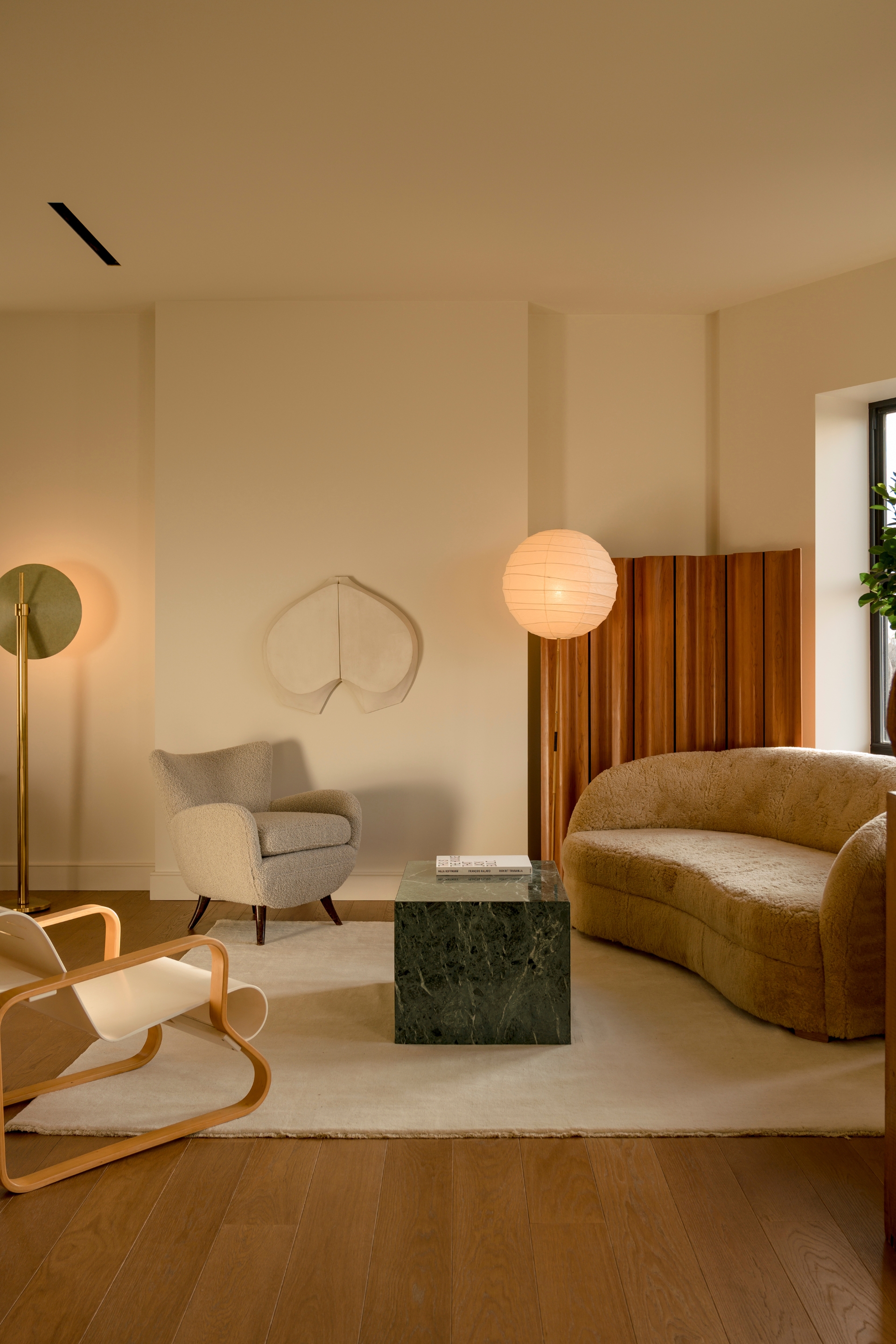 Image of a neutral living room with off-white walls and an off-white rug on top of hardwood floors. There is a curved, beige, boucle sofa, a green marble coffee table, a lantern light, and two accent chairs in the room.