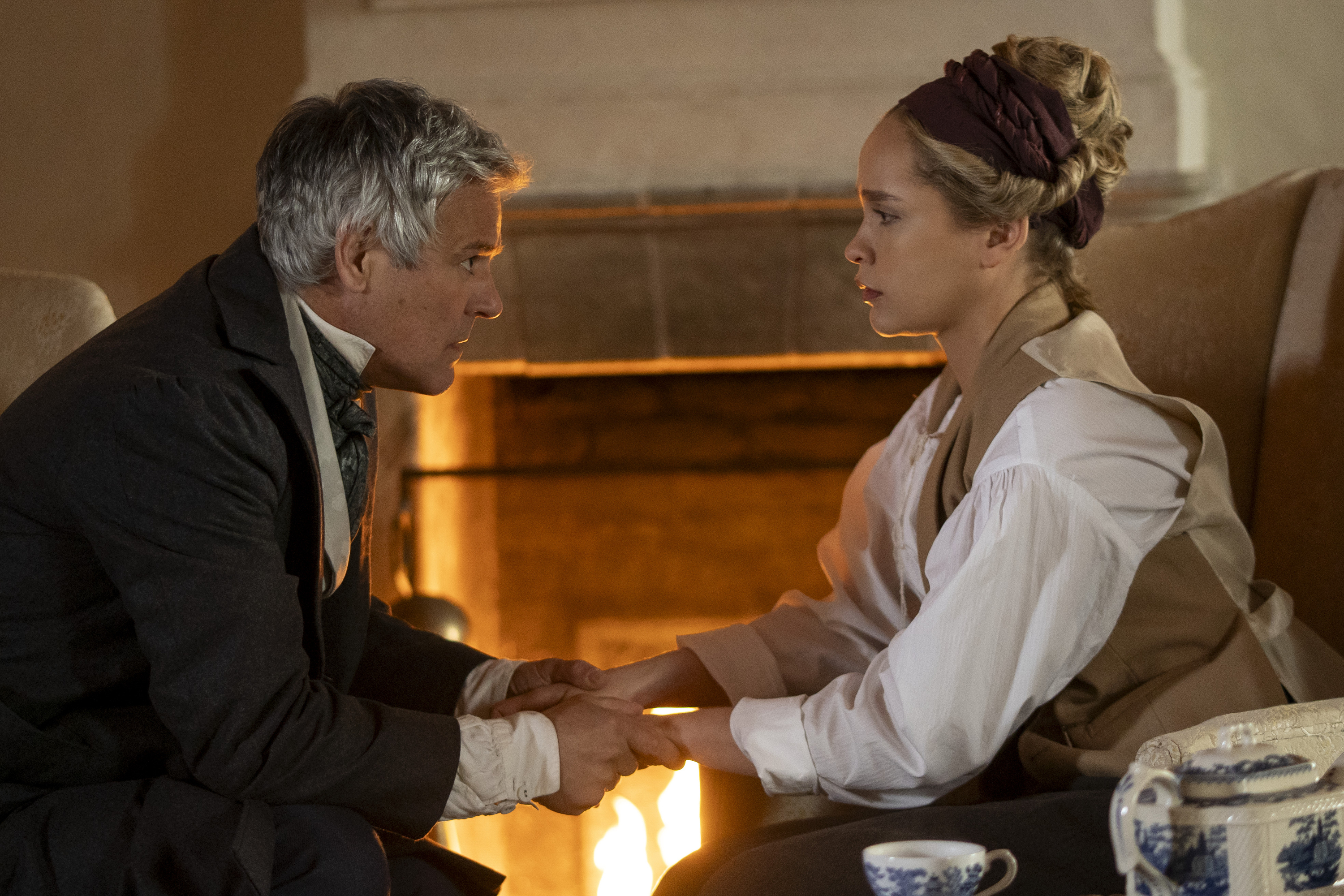 A man (Rupert Graves as Mr. Goff) and his daughter (Iola Evans as Tanna) hold hands while sitting in front of a fireplace, in 'Washington Black.'