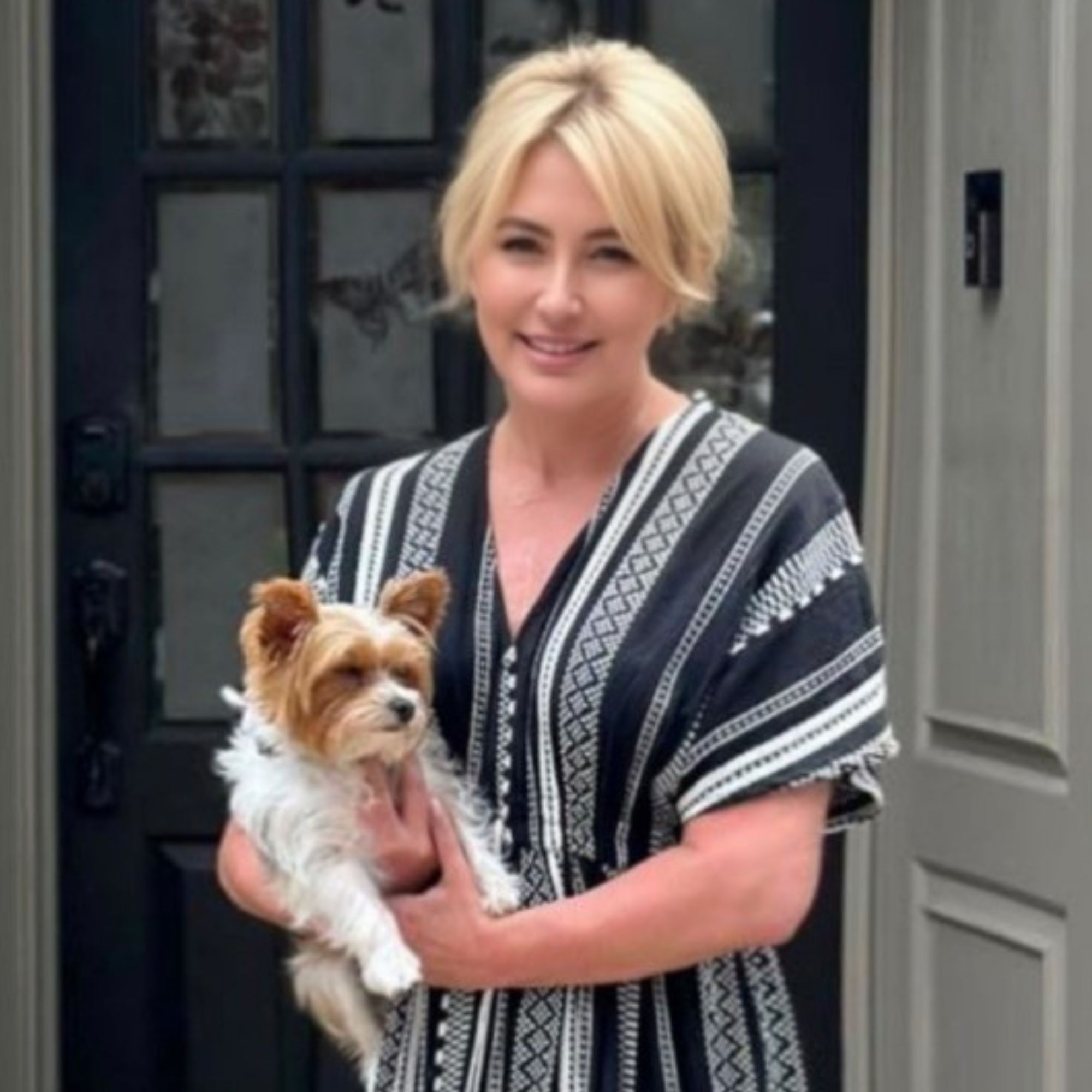 A picture of Kim Riley, a woman with tied back blonde hair wearing a striped blue, gray and white dress holding a brown and white dog, standing in front of a black door with glass rectangular panels