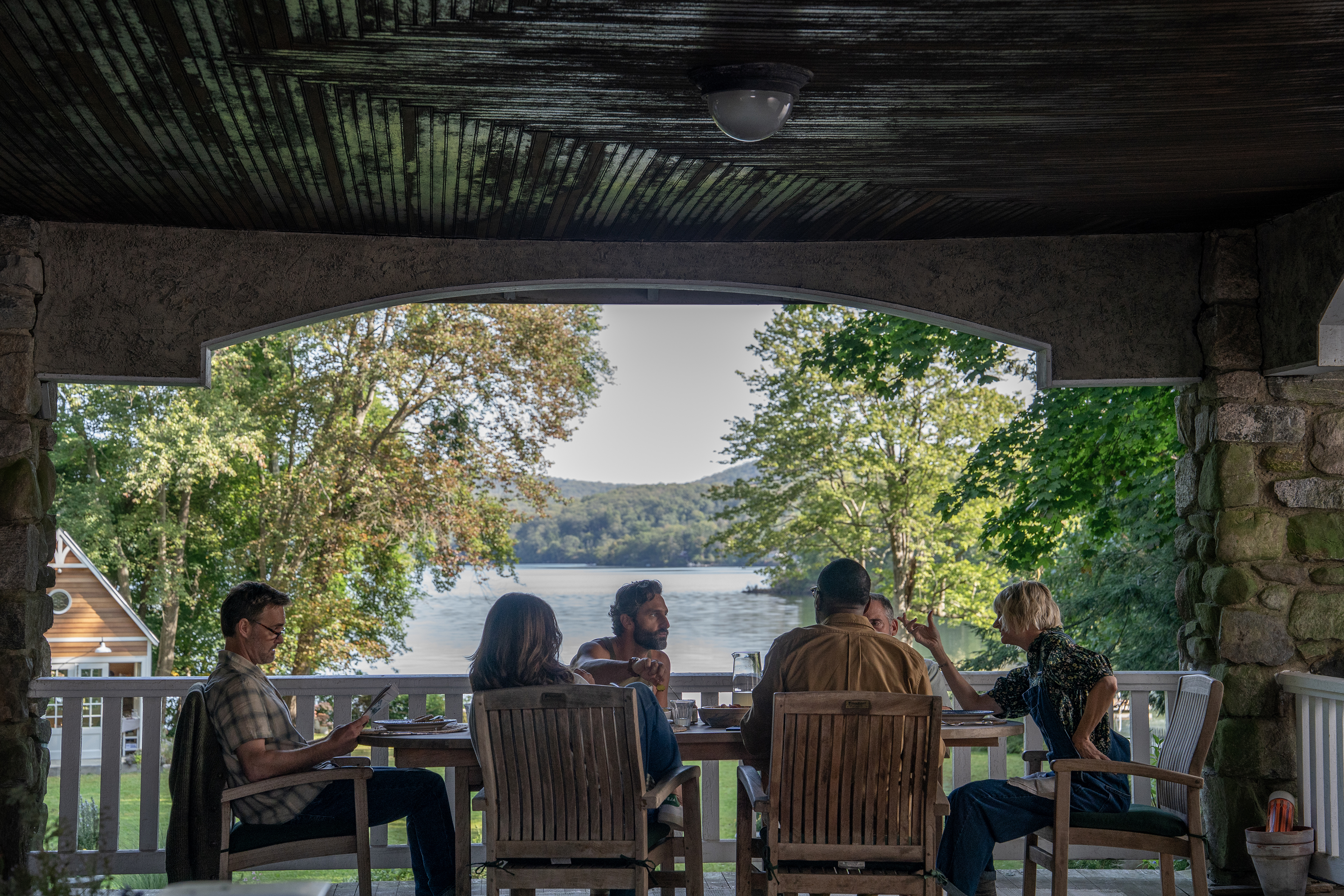 A group of people (l-r: Will Forte as Jack, Tina Fey as Kate, Marco Calvani as Claude, Colman Domingo as Danny, Steve Carell as Nick, and Kerri Kenney as Anne) sit at a dining table on a covered patio, with a lake in the background, in Episode 101 of The Four Seasons.