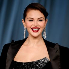 a woman with dark hair wearing a black blazer and dangly earrings and posing in front of a blue backdrop