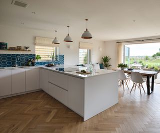 A kitchen with white units and a wooden herringbone patterned floor