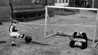 Black and white photo of a small boy kicking a ball into a football net as a panda sits, acting as goalkeeper.