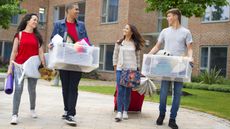 Four college students carry totes as they walk outside a dorm.