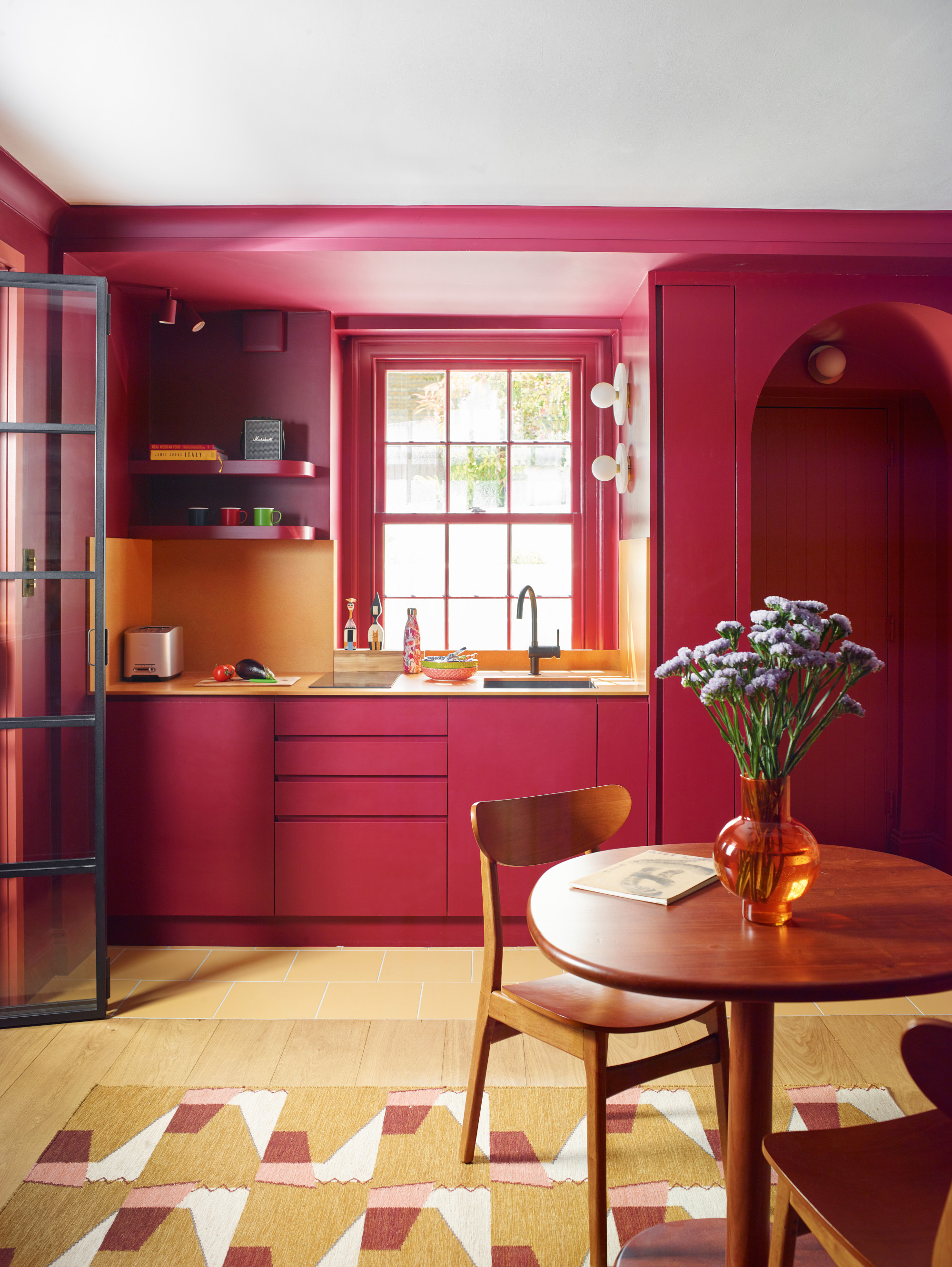 Modern kitchen cabinets in deep red with yellow countertop and island, yellow tiled floor, yellow and pink rug, wood round breakfast table and chairs, and amber glass vase