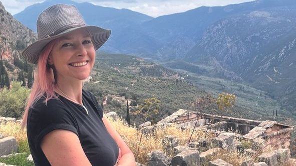 Alice Roberts in a black t-shirt and grey hat standing in front of a hilly background 