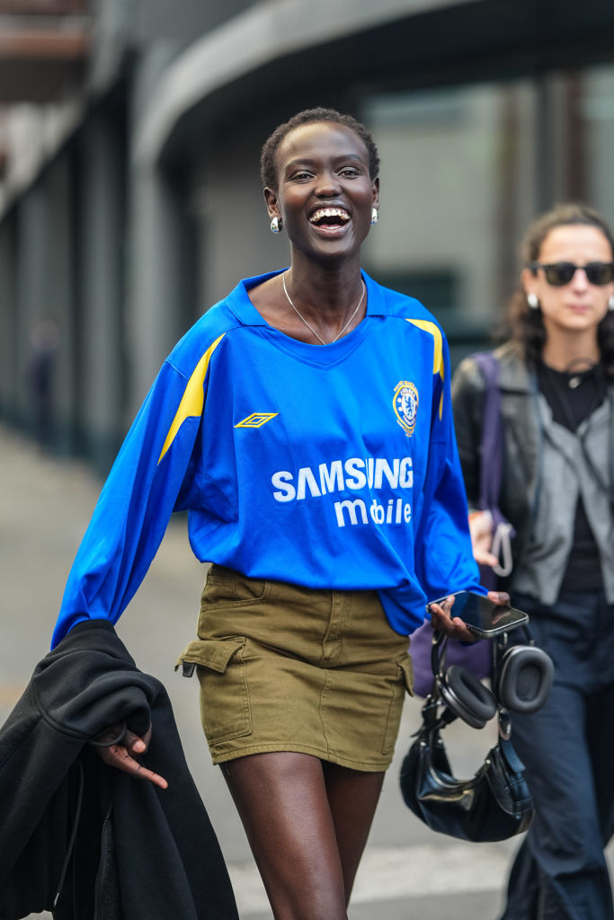 A model wears silver chrome earrings, silver necklace, bright blue oversized graphic print sports shirt, dark green olive denim jean mini skirt, shiny black leather bag, black hoodie,
