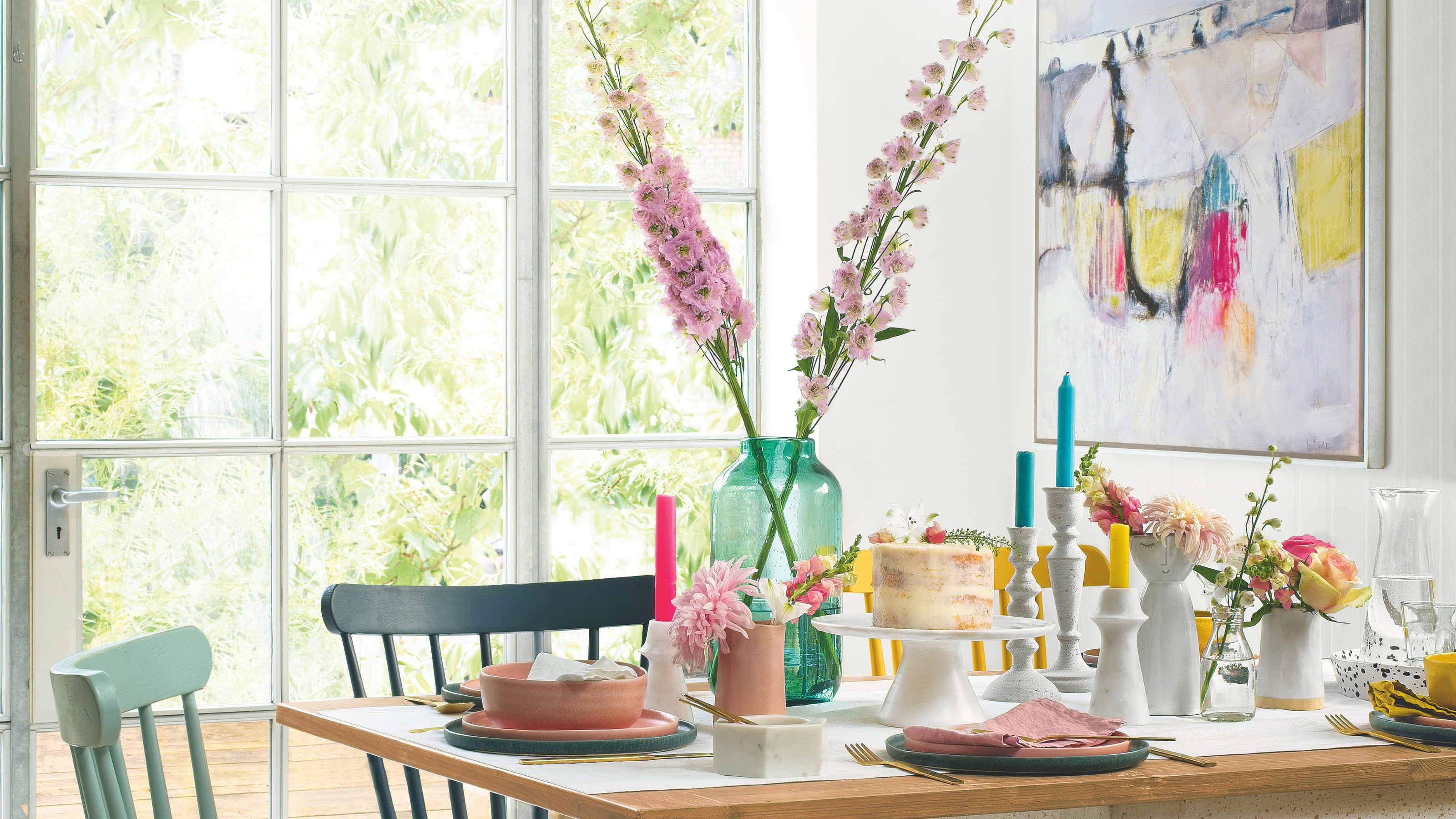 Spring tablescape with fresh pink flowers in green vase, primary coloured canles and a sponge cake on white stand.