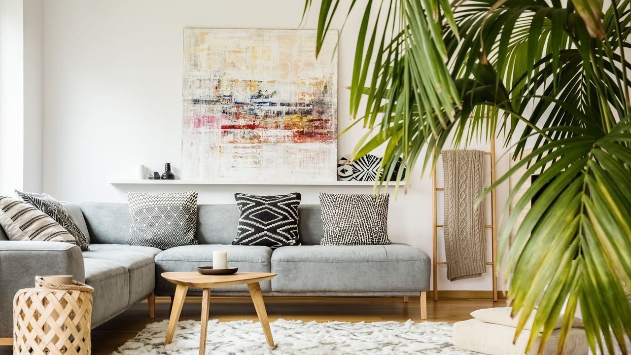 An image of a living room with a gray L-shaped sofa with monochrome throw cushions, a small triangular coffee table, a white and black rug, a shelf with a large piece of wall art, and a large house plant