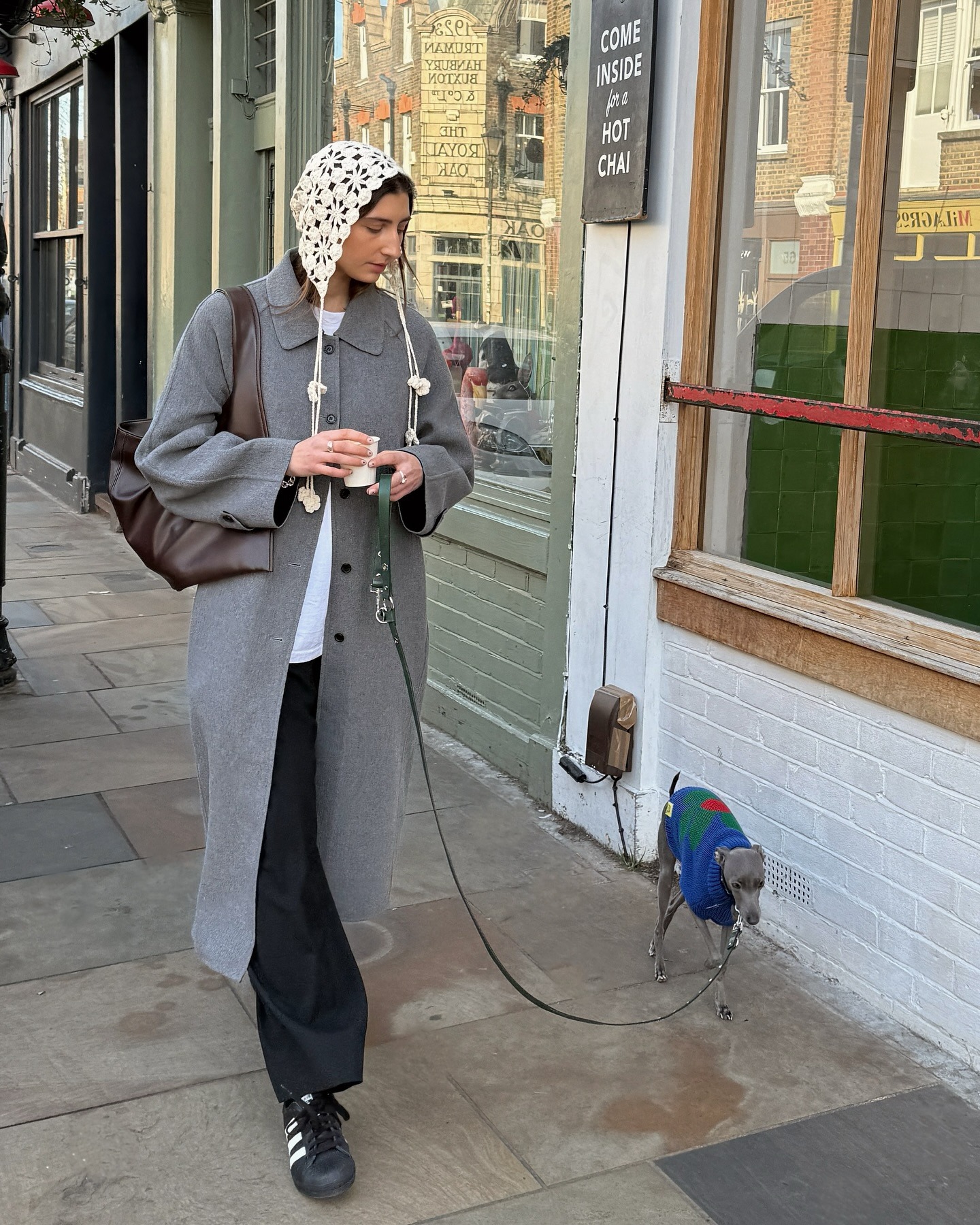 Influencer Poppy wears a crochet headscarf, a heather grey wool coat, black trousers, a slouchy, oversized leather hobo bag, and black Adidas Sambas.