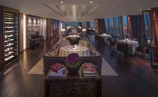 A restaurant area in the Shangri-La at the Shard hotel. To the left, we see vine fridges full of bottles. In the center, we have a row of round tables with light and dark brown chairs and couches with lamps beside them. To the right, against the floor-to-ceiling windows that overlook the city at night, we have square tables with brown chairs.