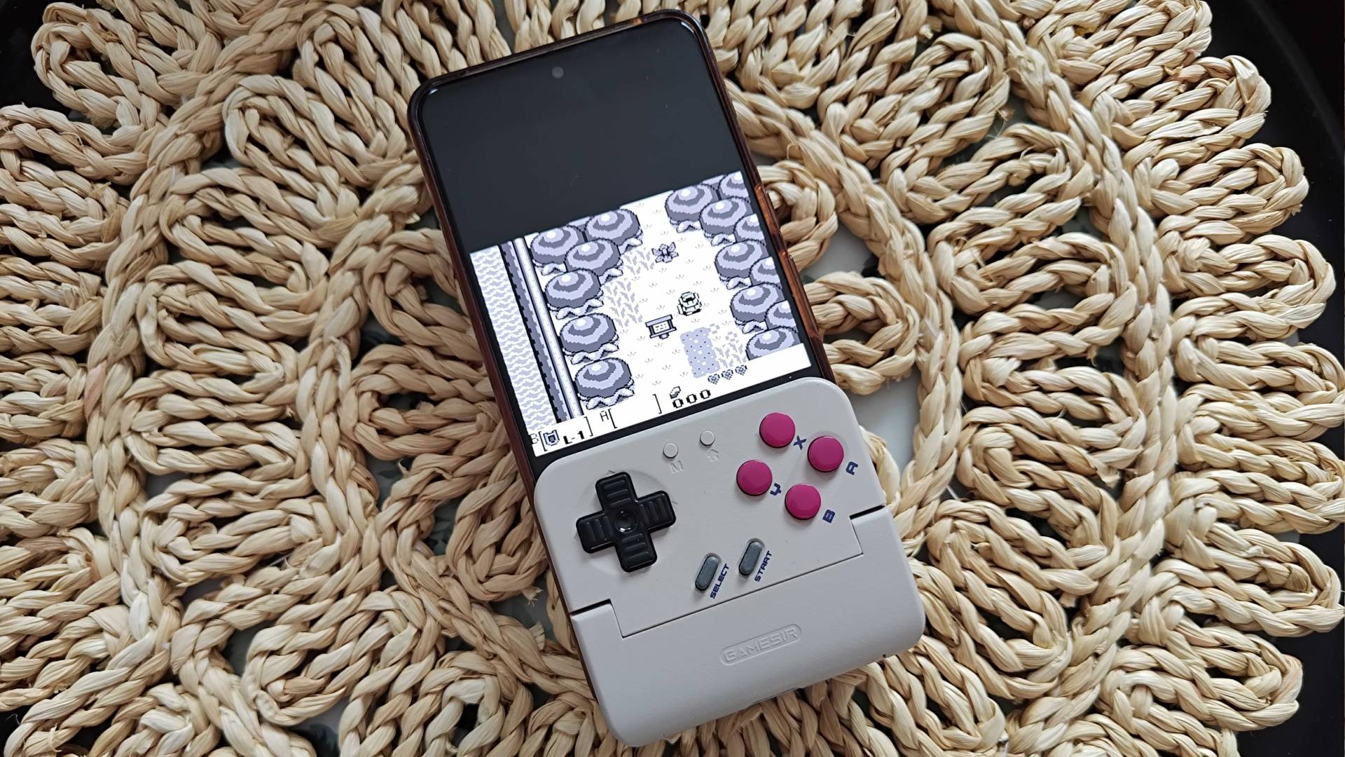 8Bitdo FlipPad on wicker table cover with Zelda: Link's Awakening gameplay on phone screen.