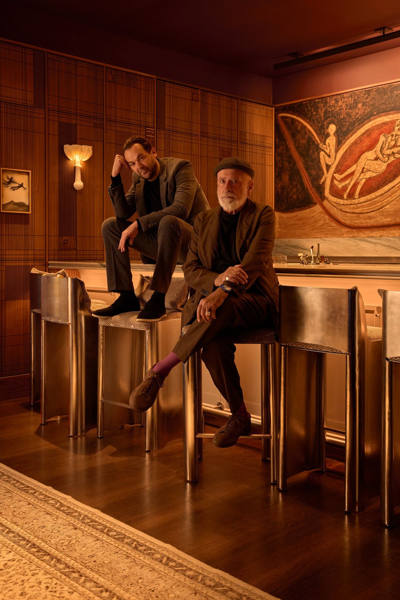 Two men dressed in dark brown suites sit in a creatively decorated bar featuring chrome tall stools, an ornate pale rug, sleek wooden floors and paneled walls, and a large-scale mythological painted mural.