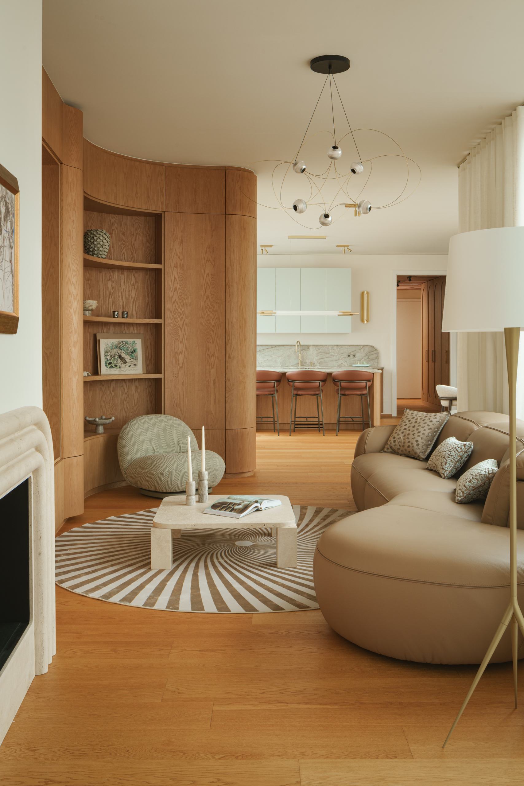 Image of a modern living room that opens to a kitchen. The living room has wooden floors and walls, a beige leather bean-shaped sofa, a spiral rug, and a white marble coffee table.