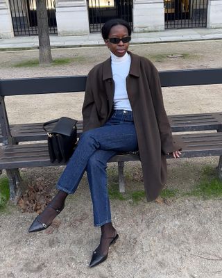 style influencer and creative Sylvie Mus sitting on a bench in Paris wearing a lightweight white turtleneck sweater, brown coat, dark-wash straight-leg jeans, black tights, and black kitten heels