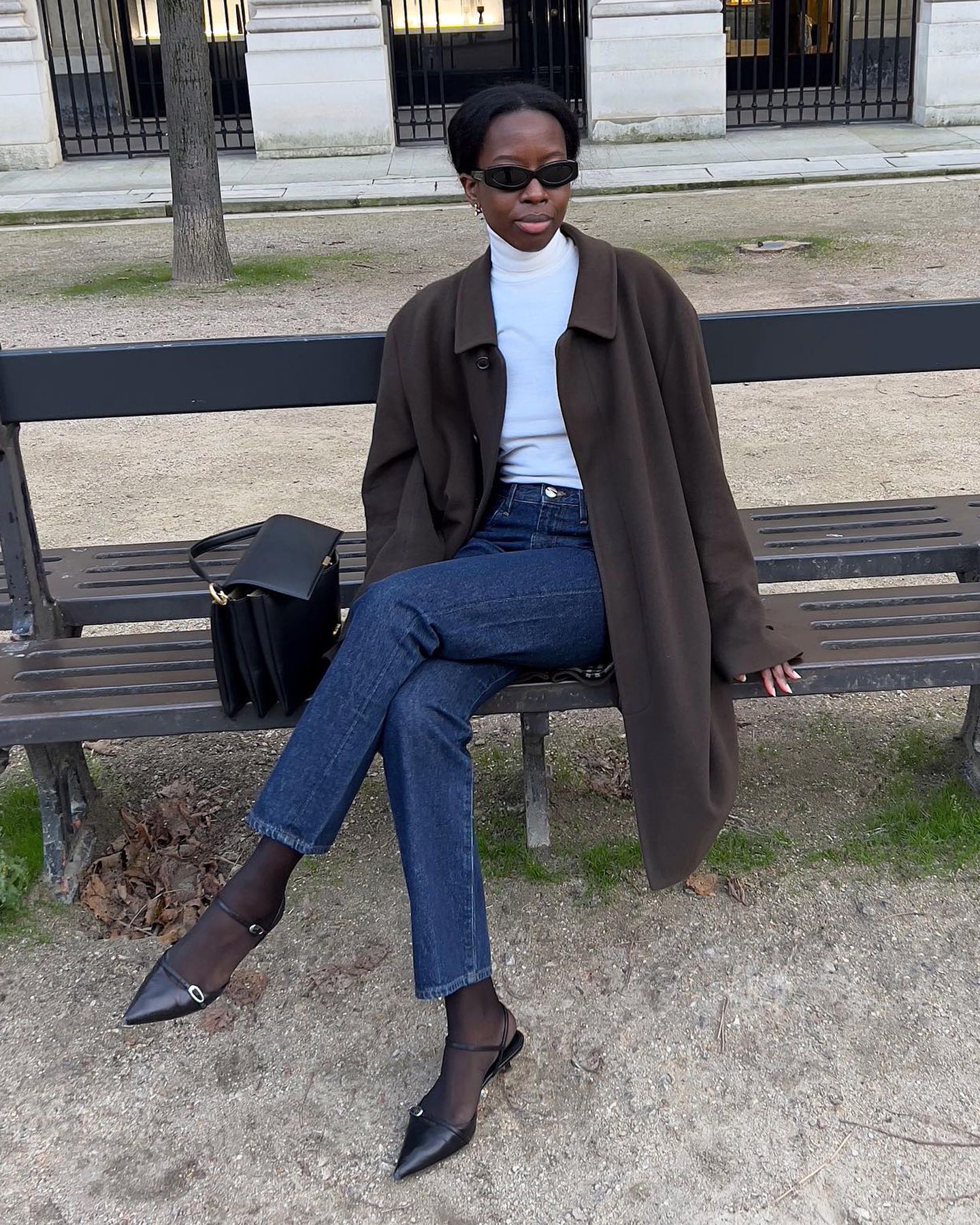 style influencer and creative Sylvie Mus  sitting on a bench in Paris wearing a lightweight white turtleneck sweater, brown coat, dark-wash straight-leg jeans, black tights, and black kitten heels