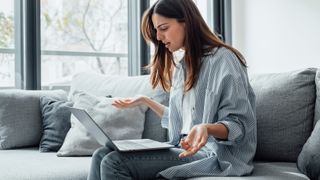 A woman sitting on a couch with a laptop and holding her hands out in confusion