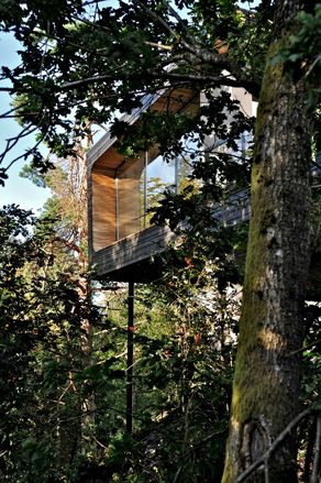 Closer exterior view of the elevated Trekronekabin - a dark wood house surrounded by trees under a blue sky