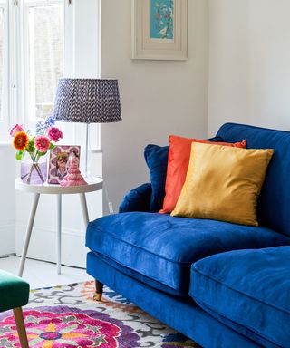 A living area with a navy blue velvet couch with yellow and orange throw pillows, white walls, a side table with flowers and a lamp, and a white, purple, and pink statement rug