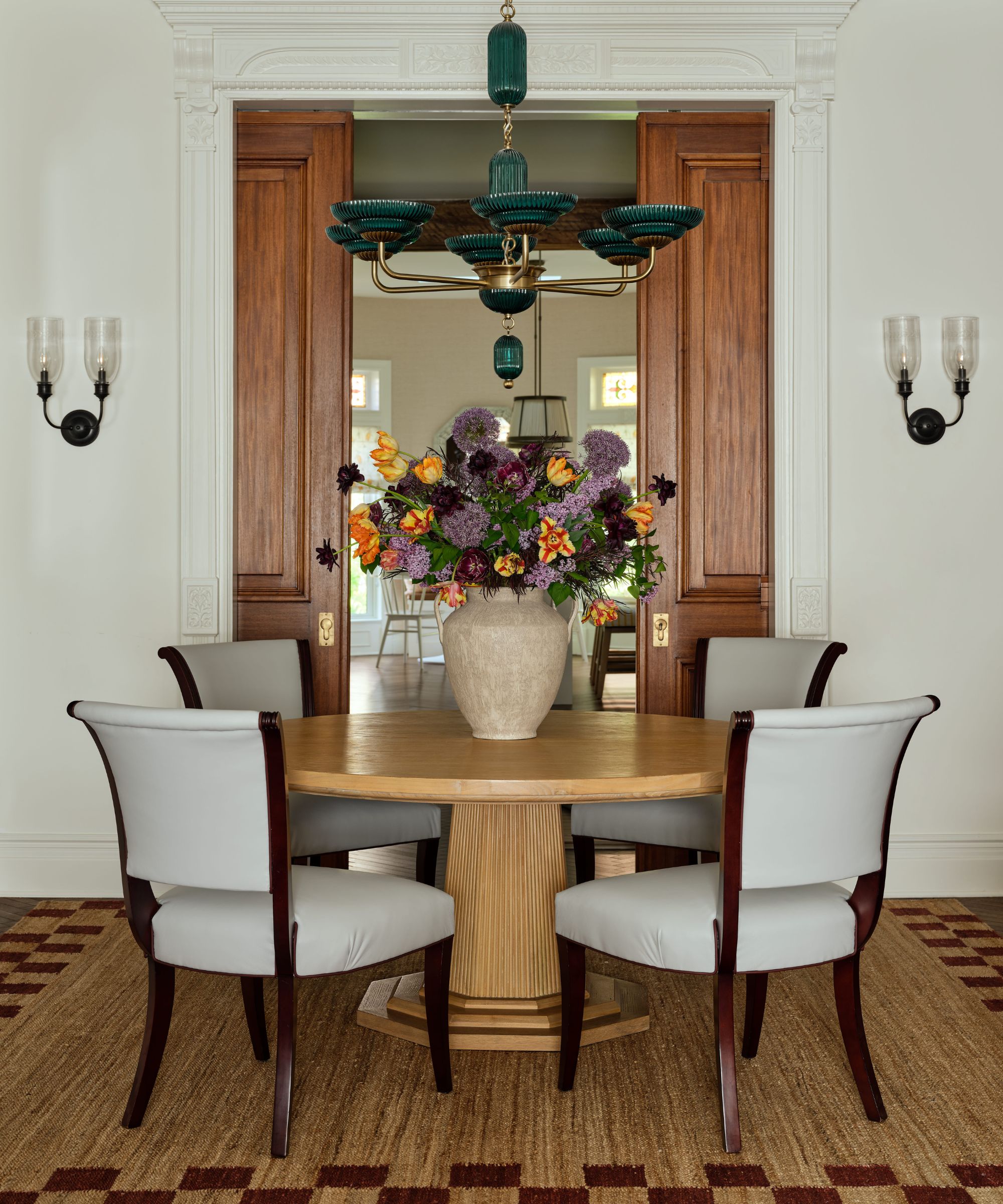 a New York brownstone dining room with original molding and pocket doors, round wooden table, antique chairs, a woven and checkerboard trimmed rug with a green vintage chandelier