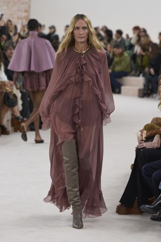 A model at Chloé's Fall/Winter 2024 show wearing a sheer, ruffled maxi dress, thigh-high boots and a gold necklace.