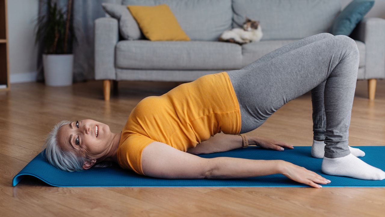 Woman in yellow T-shirt and grey leggings performing bridge exercise in domestic setting