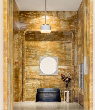 A passage with brown marble walls, a black console, a round mirror and a vase with flowers in on a round side table.