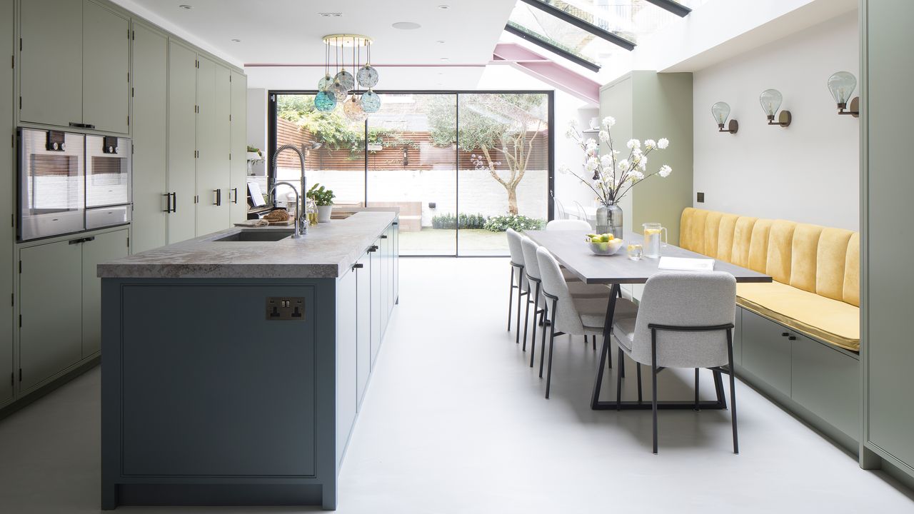 A wide shot of modern kitchen diner in Victorian Terrace house with olive green kitchen cabinetry, blue kitchen island, light grey dining set and yellow banquette seating