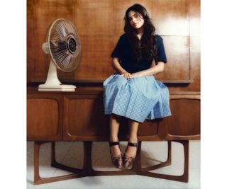 Ariana Greenblatt sitting on table wearing blue top, checkered skirt, and satin ballet flats.