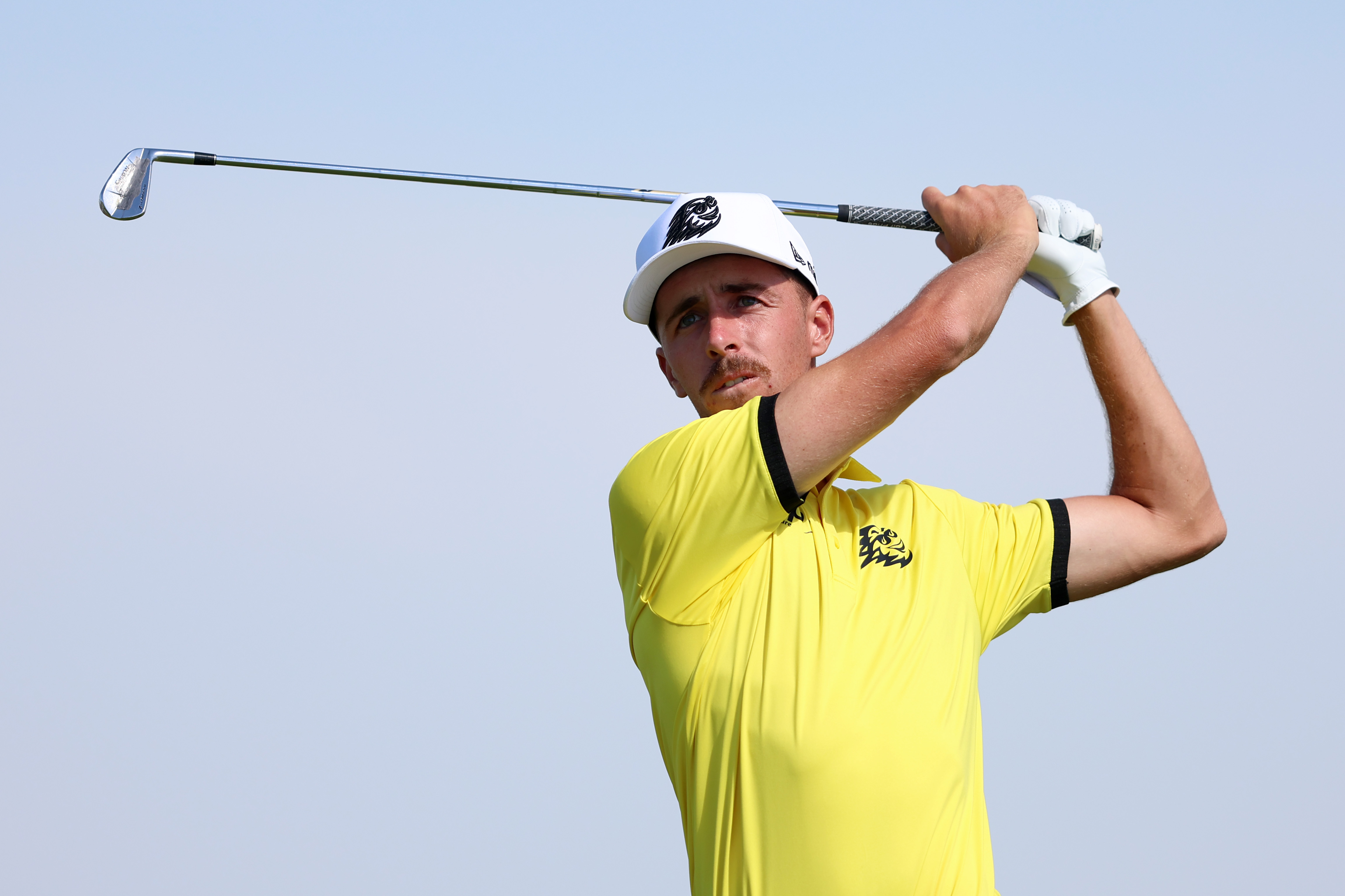 David Puig hits an iron shot wearing a yellow polo and white cap