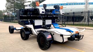 a six-wheeled, blue-and-white moon rover sits on a sidewalk in front of a large white airplane