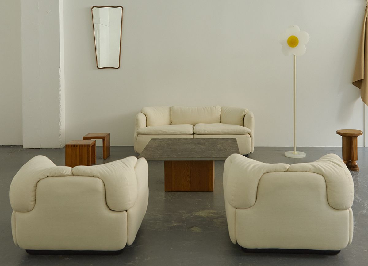In an industrial gallery, three cream couches, a wood and stone coffee table, a couple of wooden stools, a mirror and a daisy-shaped lamp stand in the same room recreating a living room setting.