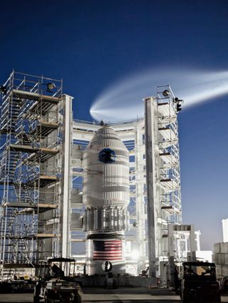 A white conical vessel with round window near the top stands surrounded by white scaffolding under a dark blue evning sky. Above, a giant, jelly-fish like plume trails a rocket launching through the sky.