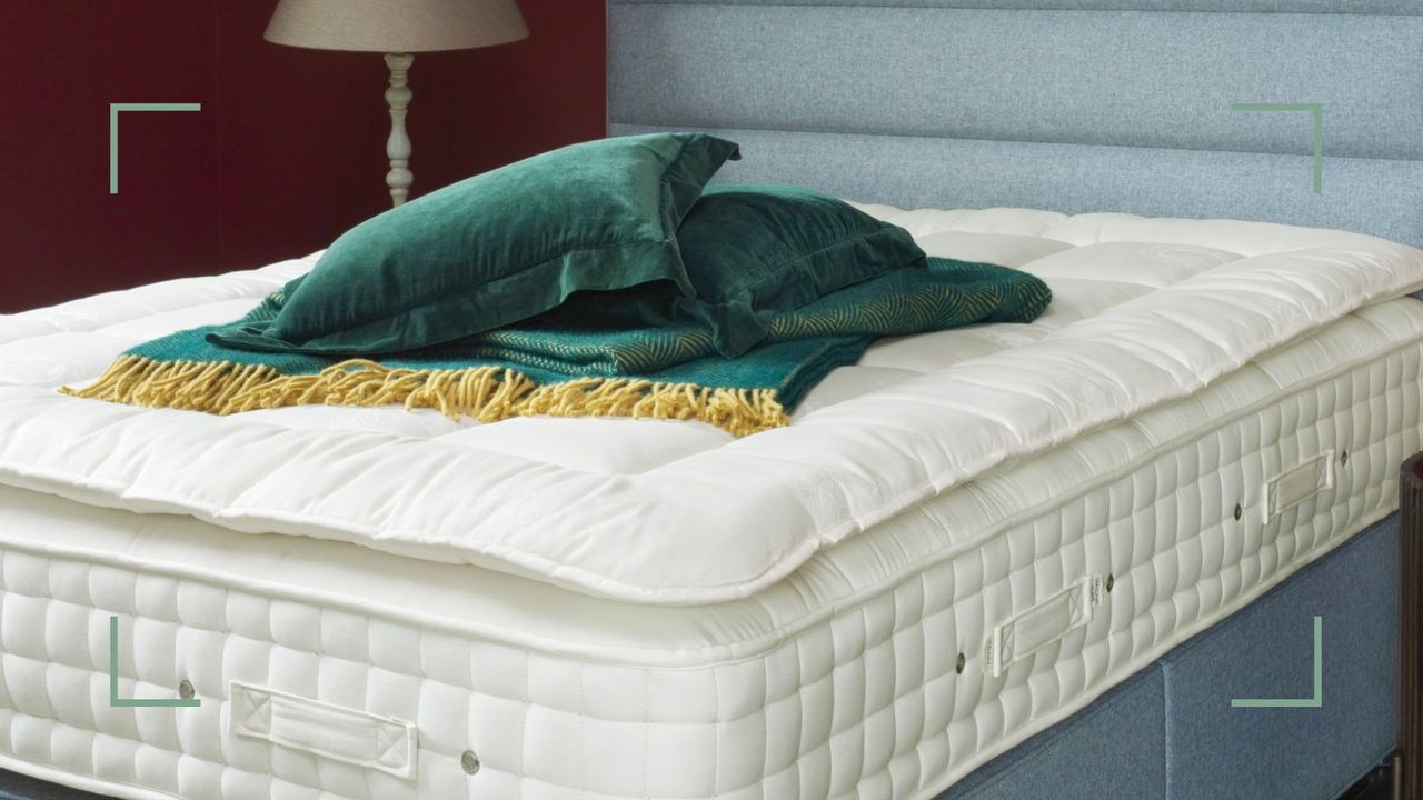 Hypnos Beds mattress topper on a bed with green blankets and pillows on top, in a bedroom with a red wall and a beige lamp, to illustrate how to keep a mattress topper from sliding