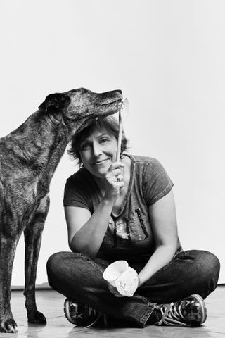 A dog is licking an ice cream spoon held by a Elke sitting cross-legged on the floor