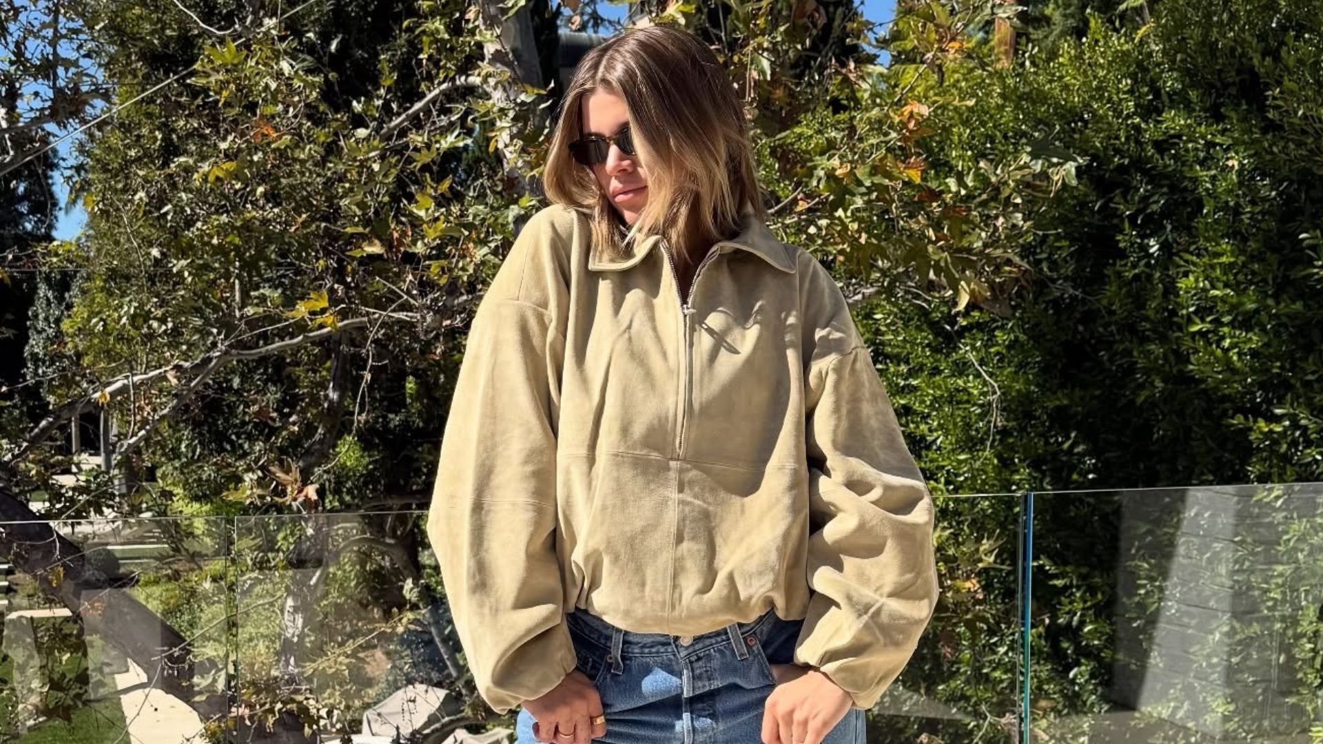 Sofia Richie Grainge poses on a balcony wearing a light beige suede jacket, blue vintage Levi jeans and satin pointe-toe heels. She holds a black leather bag in her hand and wear black sunglasses.