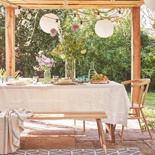 outdoor dining garden party table setting, linen tablecloth, flowers on table, bench, blanket, rug, pergola