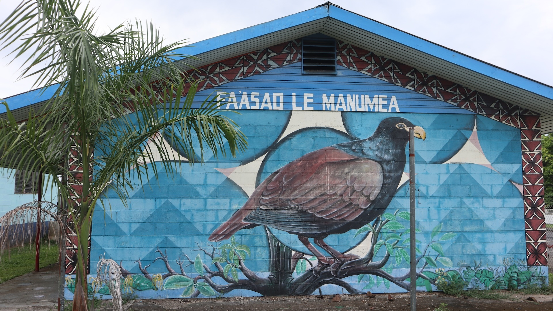 Photo of a the exterior of a building on which is a mural of a large bird as viewed from the side. Above the bird it says, "PAASAO LE MANUMEA."