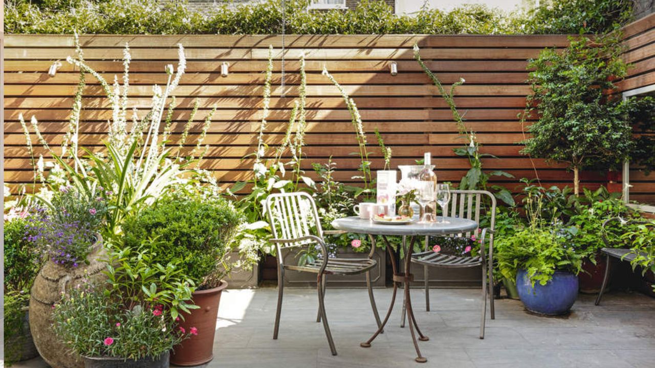 Garden patio with wood panelled walls and a small bistro set, surrounded by potted plants