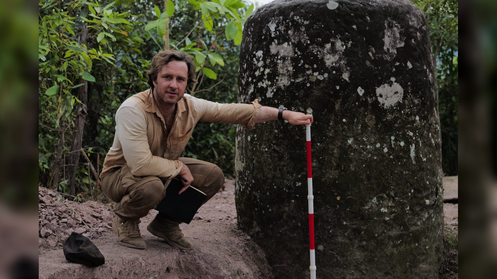 a light-skinned man in khaki clothing squats next to a tall stone jar with a meter stick