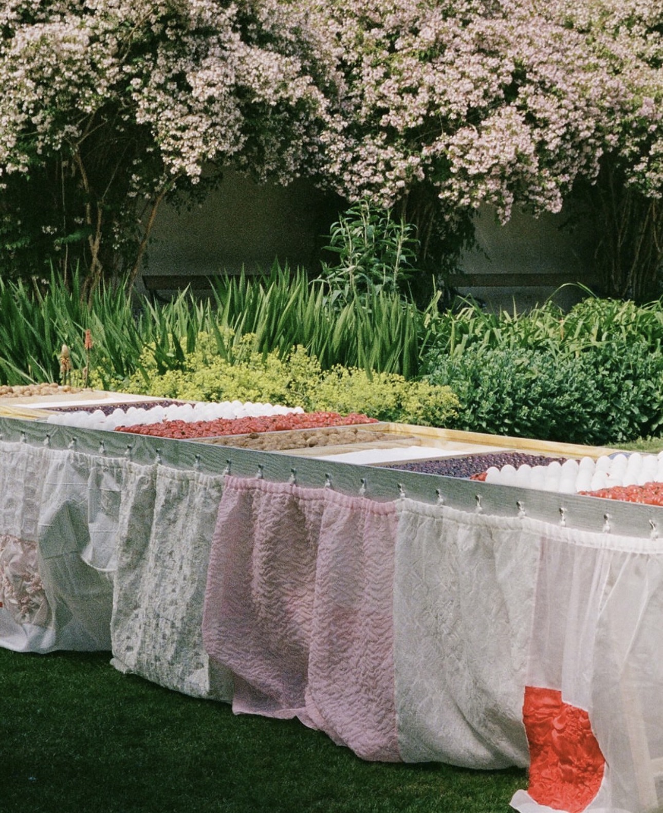 A culinary garden pop-up with a quilted table in soft, romantic fabrics in white and pink, with a surface divided into rectangulars filled with different foods, like cherries, tomatoes, and eggs.