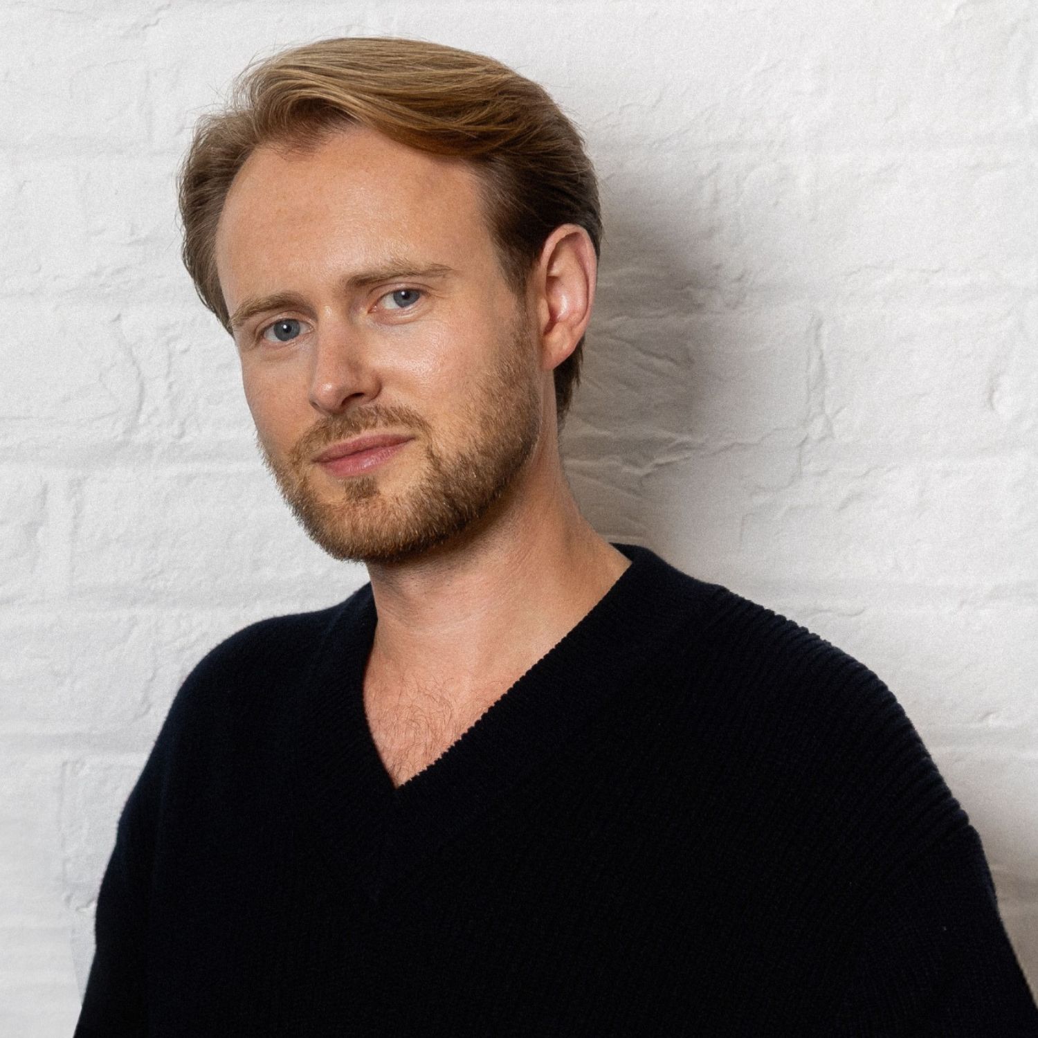 A man with short brown hair in a black v-neck in front of a white brick wall