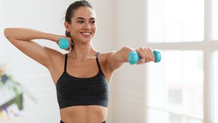 Woman holding two light dumbbells during workout with left arm extended in front of her and right arm bent