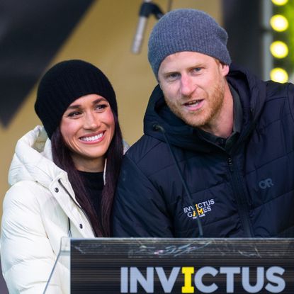 Meghan Markle wearing a white coat and black beanie smiling next to Prince Harry who is speaking at a podium wearing an Invictus Games jacket and waving