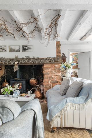 living room with white and gray sofas and fireplace with wood burner and vintage copper pots and white painted beamed ceiling with twig decoration