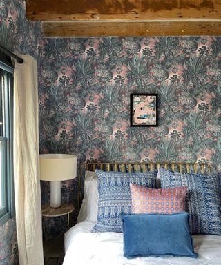 farmhouse style bedroom with blue and pink wallpaper, beams, blue and pink bed pillows, table lamp, cream drapes