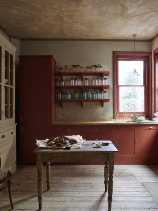 Rust red kitchen cabinetry, with red-painted window frame, rustic wooden floorboards and dining table and open shelving.