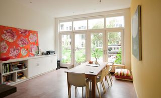 Room with white low cabinet against a white wall with orange floral wall art. o the left id dinig set (table and chairs) next to a yellow wall. The room has white french doors leading outside