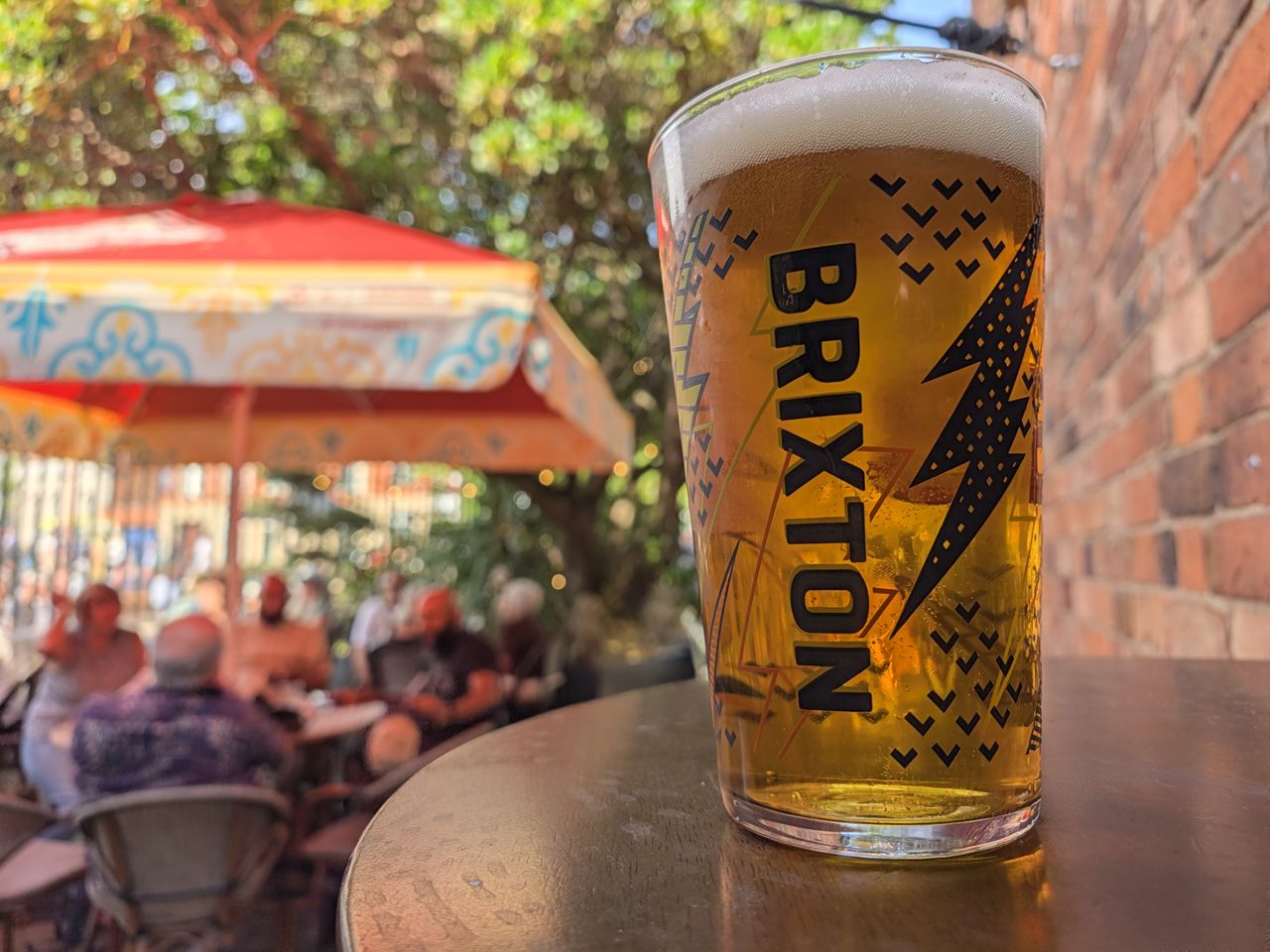 Close-up of a pint of Brixton beer on an outdoor pub table, with people and colorful umbrellas blurred in the background.